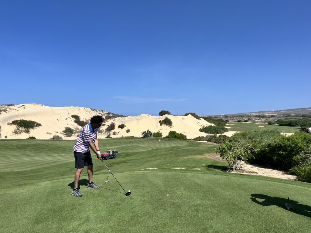 Player teeing off at Diamante Dunes with sand dunes behind