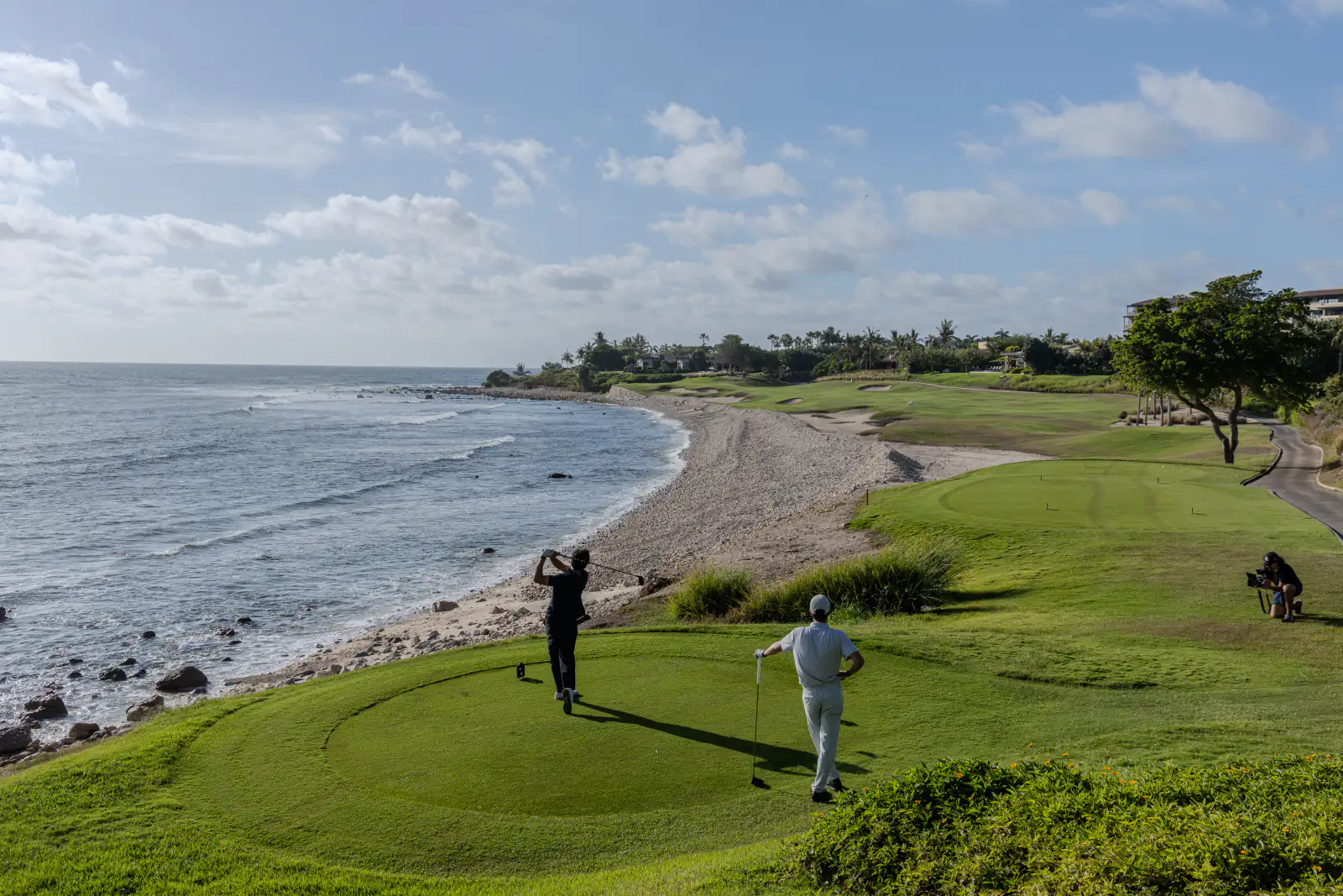 Two golfers on a coastal tee at Punta Mita, with the beach curving away behind them and the Pacific to the left.