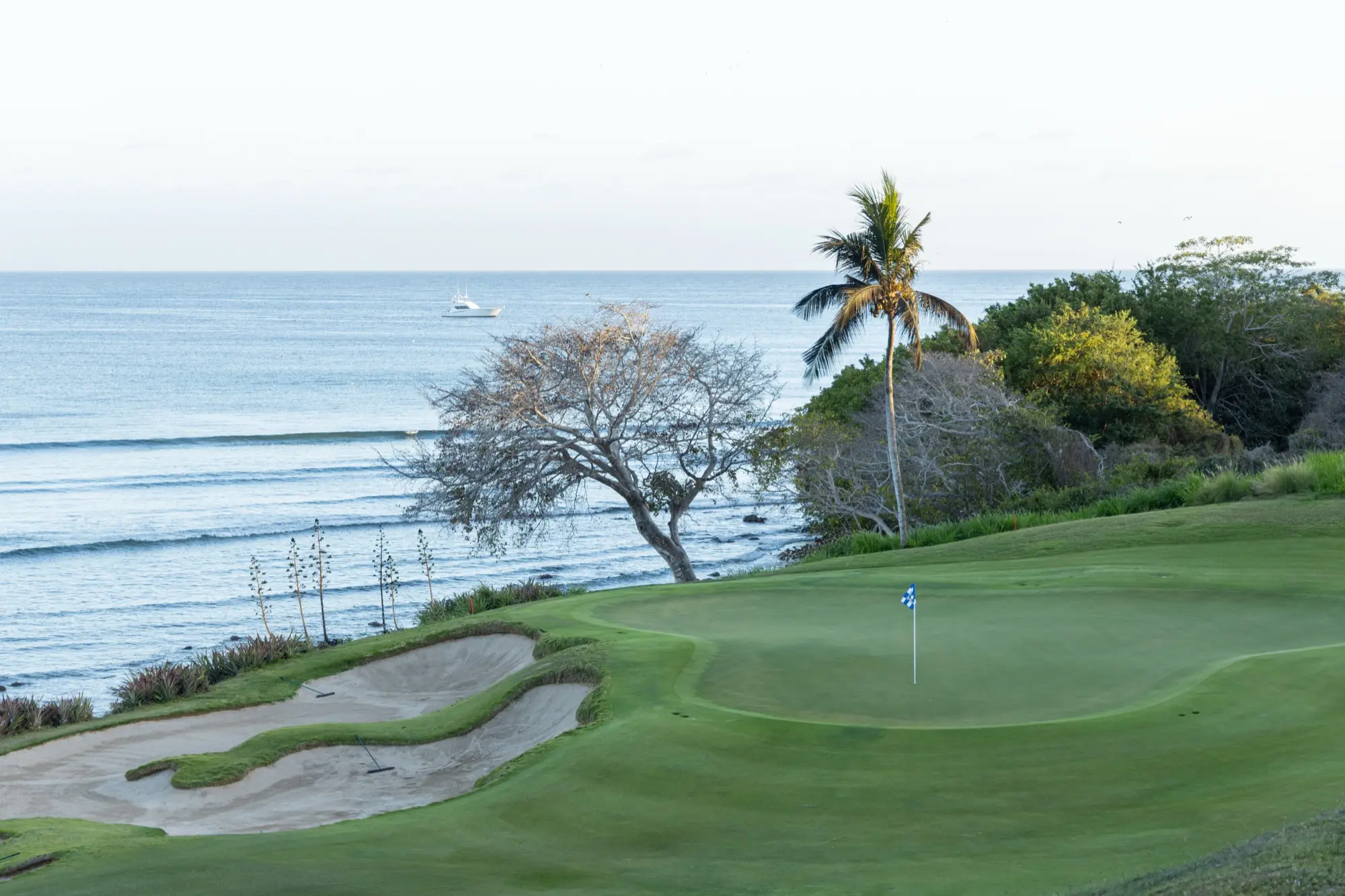 A green and flagstick on the Punta Mita peninsula with a bunker short and surf breaking beyond.