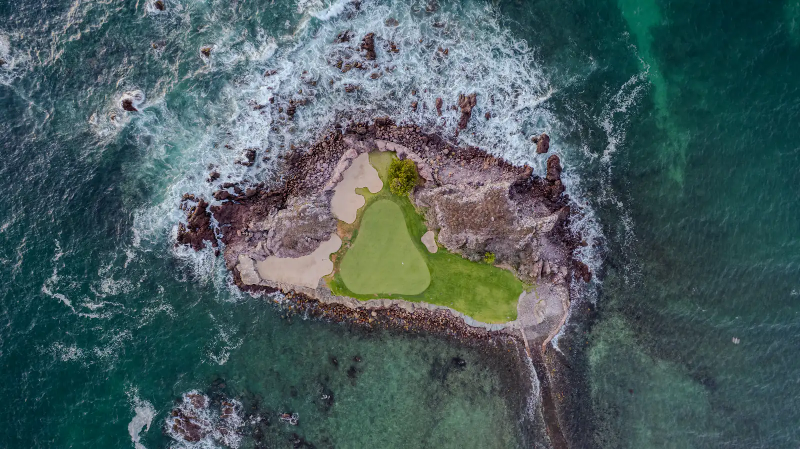 Aerial view of the Tail of the Whale, hole 3B at Pacifico Course in Punta Mita — a natural island green ringed by surf, with the stone causeway visible at low tide.