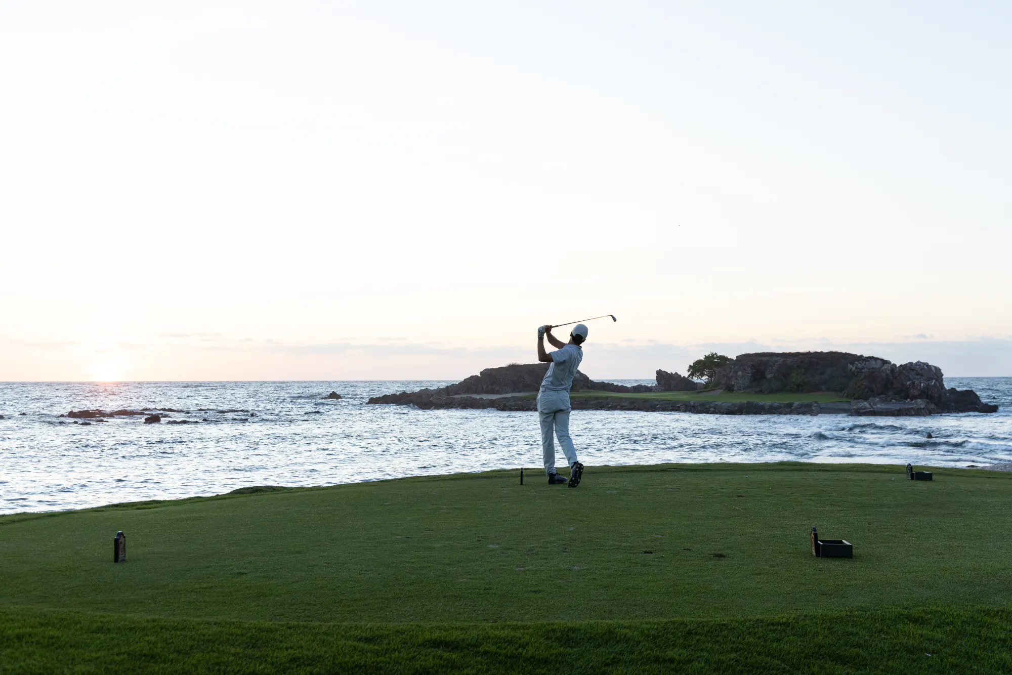 A golfer mid-swing at Pacifico Course, Punta Mita, playing toward the island green at hole 3B with the Pacific behind.